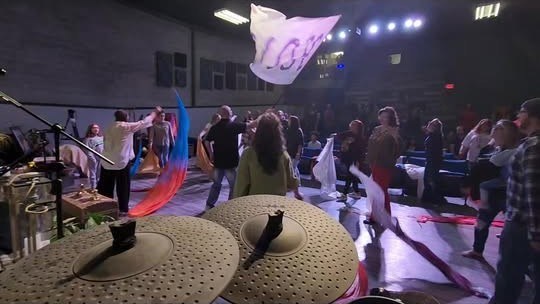 A worship gathering seen from behind the drums, with flags in the room.
